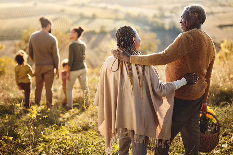A grandfather puts his arm around his granddaughter with family in the background. A mother, father, and young daughter smile while engaging in a happy group hug.