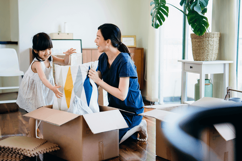 Mother unpacks a box of colorful throw pillows with her young daughter in their new home.