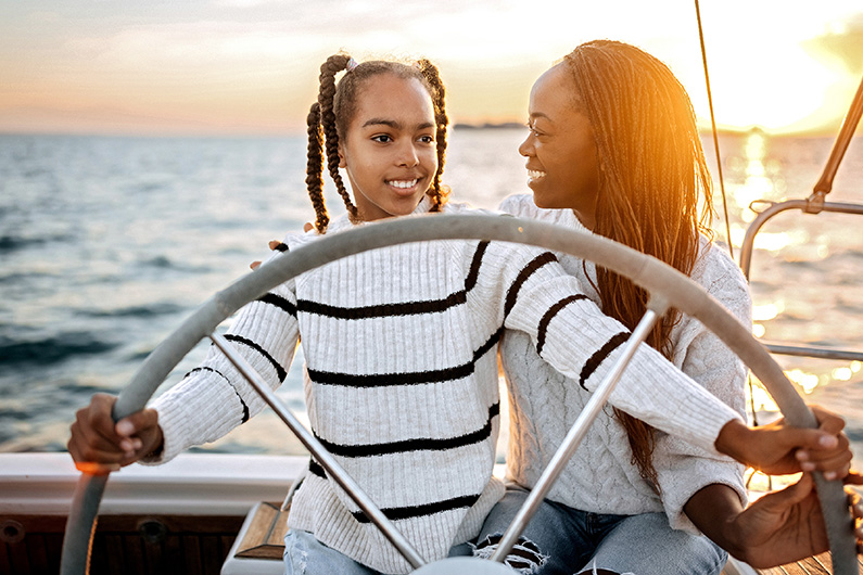 Two people on a boat at sunset, one person is steering the boat while the other sits beside them. Both are wearing white sweaters with black stripes.