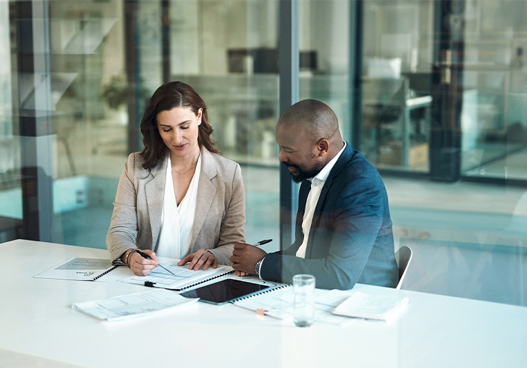 A man and a woman sitting at a desk in an office, discussing documents and taking notes.